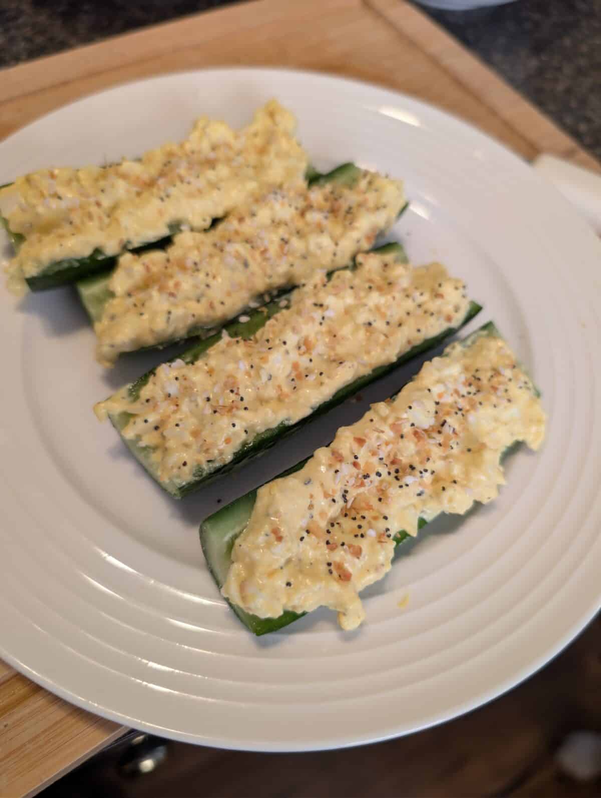 Four cucumber halves on a white plate, these Egg Salad Cucumber Boats are filled with a creamy egg mixture and sprinkled with seasonings for a refreshing, tasty bite.