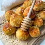 A pile of golden baked pastry balls on a wooden board, drizzled with honey using a honey dipper and sprinkled with crushed nuts, evokes the delightful look of Honey Almond Cookies, with a white cloth in the background.