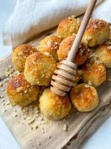 A pile of golden baked pastry balls on a wooden board, drizzled with honey using a honey dipper and sprinkled with crushed nuts, evokes the delightful look of Honey Almond Cookies, with a white cloth in the background.