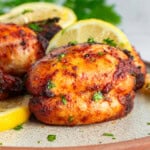 Close-up of grilled chicken thighs garnished with chopped parsley and lemon slices on a speckled plate. The chicken appears golden and well-seasoned, with a backdrop of fresh parsley leaves.