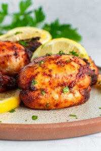 Close-up of grilled chicken thighs garnished with chopped parsley and lemon slices on a speckled plate. The chicken appears golden and well-seasoned, with a backdrop of fresh parsley leaves.