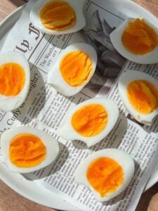 Several hard-boiled eggs, sliced in half to reveal their bright orange yolks, are arranged on a white plate lined with a sheet of newspaper. Sunlight casts shadows across the eggs and newspaper.