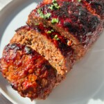 Air Fryer Meatloaf sliced and topped with a caramelized, glossy glaze, garnished with chopped herbs, served on a round white plate in bright sunlight.