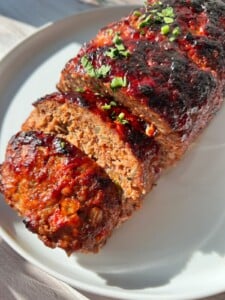 Air Fryer Meatloaf sliced and topped with a caramelized, glossy glaze, garnished with chopped herbs, served on a round white plate in bright sunlight.