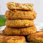 A stack of golden-brown breaded patties, some showing a creamy inside with herbs. A partially eaten patty is on top, revealing a soft filling. Fresh green leaves are scattered around on a white surface.