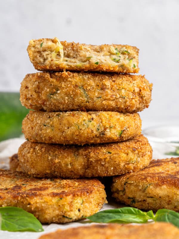 A stack of golden-brown breaded patties, some showing a creamy inside with herbs. A partially eaten patty is on top, revealing a soft filling. Fresh green leaves are scattered around on a white surface.