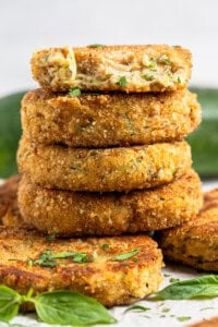 A tall stack of crispy, golden-brown zucchini fritters with flecks of green herbs. One fritter has a bite taken out, revealing a moist interior. Basil leaves are scattered around the base of the stack.