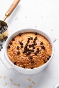 A baked oatmeal dish in a white bowl, topped with chocolate chips, sits on a light surface. Nearby are scattered oats and a brass measuring cup filled with more chocolate chips. The dish has a golden-brown appearance.