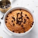 A white bowl filled with baked oatmeal topped with chocolate chips sits on a marble surface. Nearby is a brass measuring cup with more chocolate chips scattered around. Some oats are visible on the surface.