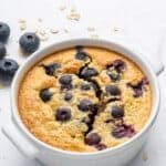 A baked oat dish topped with blueberries, served in a white ramekin. The surface is golden brown with bursts of juicy blueberries. Some oats and fresh blueberries are scattered on the white background.