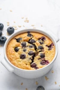 A baked oat dish topped with blueberries, served in a white ramekin. The surface is golden brown with bursts of juicy blueberries. Some oats and fresh blueberries are scattered on the white background.