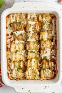 A white baking dish filled with cheesy Eggplant Rolls, topped with melted cheese and chopped parsley, sits on a white surface. The neatly arranged rolls rest atop a rich tomato sauce, making for an inviting presentation.