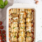 Baked Eggplant Rolls rollatini in a white dish, topped with melted cheese and herbs, surrounded by fresh cherry tomatoes, basil leaves, garlic cloves, salt, and a gray napkin on a marble surface.