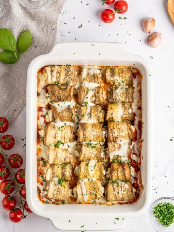 Baked Eggplant Rolls rollatini in a white dish, topped with melted cheese and herbs, surrounded by fresh cherry tomatoes, basil leaves, garlic cloves, salt, and a gray napkin on a marble surface.