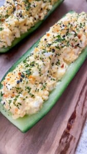 A cucumber half is hollowed out and filled with a creamy egg salad mixture, garnished with herbs and seasonings, and placed on a wooden board.