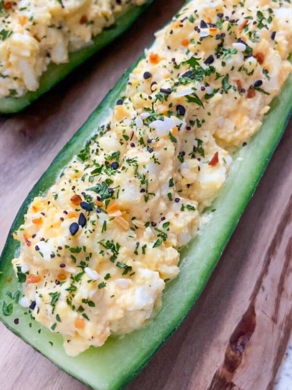 Egg Salad Cucumber Boats feature a halved cucumber filled with creamy egg salad, topped with chopped herbs and seasoning, and served on a wooden board.