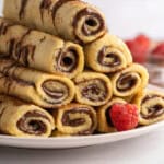 A stack of rolled crepes, reminiscent of French Toast Rollups, filled with chocolate hazelnut spread and drizzled with more chocolate, is arranged on a white plate with fresh raspberries scattered around.