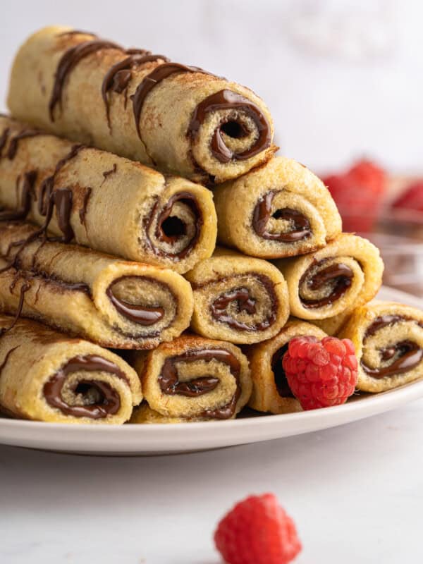 A stack of rolled crepes, reminiscent of French Toast Rollups, filled with chocolate hazelnut spread and drizzled with more chocolate, is arranged on a white plate with fresh raspberries scattered around.