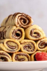 A stack of French Toast Rollups filled with chocolate and drizzled with more chocolate sits on a white plate next to a fresh raspberry.