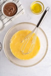 A glass bowl with whisked eggs and a metal whisk, surrounded by bowls containing olive oil, chocolate spread, and sugar—perfect ingredients for making delicious French Toast Rollups on a white countertop.