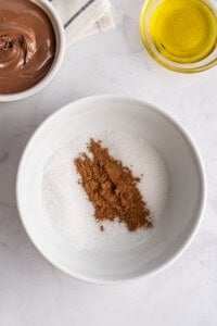 A white bowl filled with granulated sugar and cocoa powder sits on a marble surface, ready for coating delicious French Toast Rollups, alongside a bowl of chocolate spread and a small bowl of yellow oil.