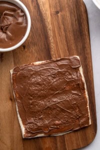 A slice of bread spread with chocolate hazelnut spread sits on a wooden cutting board, next to a small white bowl of more spread—perfect for making delicious French Toast Rollups.