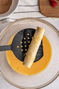 A rolled tortilla for French Toast Rollups is held over a bowl of beaten eggs with a black slotted spatula, ready to be coated. In the background, cinnamon sugar awaits alongside a wooden board topped with strawberries.
