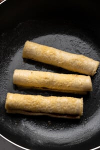 Three French Toast Rollups are being fried in a black skillet, lightly browned and sizzling in oil.