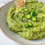 A bowl of creamy homemade guacamole garnished with sliced green chili, fresh herbs, diced red onion, and served with tortilla chips on the side.