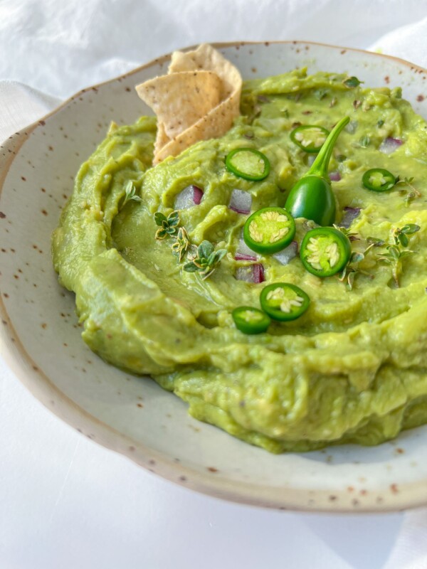 A bowl of creamy homemade guacamole garnished with sliced green chili, fresh herbs, diced red onion, and served with tortilla chips on the side.
