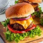 Two cheeseburgers on a wooden board with lettuce, tomato, cheddar cheese, red onion, and pickles, on a brioche bun. A striped napkin is in the background.