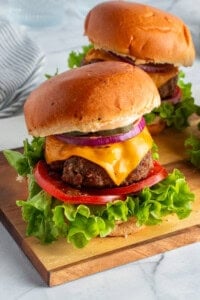 Two cheeseburgers on a wooden board with lettuce, tomato, cheddar cheese, red onion, and pickles, on a brioche bun. A striped napkin is in the background.