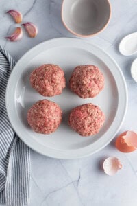 Four raw meatballs on a white plate, surrounded by garlic cloves, an empty gray bowl, a striped cloth, and cracked eggshells on a marble countertop.