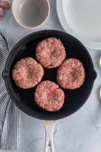 Four raw hamburger patties in a black skillet on a marble surface. Nearby are a striped cloth, garlic cloves, and empty bowls.