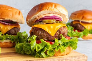Three delicious cheeseburgers with lettuce, tomato, cheese, pickles, and red onion, topped with a creamy sauce in toasted buns, are displayed on a wooden board.