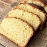 Three thick slices of golden, fluffy bread are arranged on a piece of newspaper on a wooden table, with the rest of the loaf visible in the background. The bread has a shiny, browned crust.