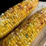 Two grilled Air Fryer Corn on the Cob sprinkled with melted butter, chopped herbs, and coarse salt, resting on a wooden surface. The corn looks golden, juicy, and delicious.