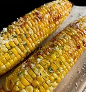 Two grilled Air Fryer Corn on the Cob sprinkled with melted butter, chopped herbs, and coarse salt, resting on a wooden surface. The corn looks golden, juicy, and delicious.