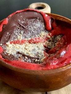 A wooden bowl filled with dark chocolate, bright red berry sauce, and a sprinkle of crushed nuts, this dessert resembles a decadent strawberry smoothie bowl with its rich flavors and vibrant presentation.