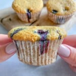 A person with light-painted nails holds an Almond Flour Blueberry Muffin in a paper wrapper, with three more muffins on a wooden board in the background. The muffin has visible blueberries and a golden-brown top.