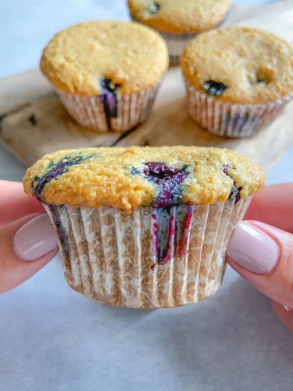 A person with light-painted nails holds an Almond Flour Blueberry Muffin in a paper wrapper, with three more muffins on a wooden board in the background. The muffin has visible blueberries and a golden-brown top.