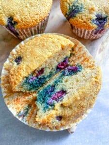 A close-up of Almond Flour Blueberry Muffins cut in half, revealing a moist interior with juicy blueberries. Whole muffins in paper liners rest on a wooden surface in the background.
