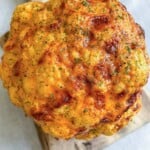 A Whole Roasted Cauliflower head with a golden-brown crust, sprinkled with herbs, rests elegantly on a wooden cutting board.