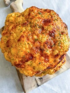 A Whole Roasted Cauliflower head with a golden-brown crust, sprinkled with herbs, rests elegantly on a wooden cutting board.