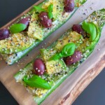 Two Egg Salad Cucumber Boats topped with sliced avocado, Kalamata olives, fresh basil leaves, hemp seeds, and seasoning, served on a wooden board against a dark background.