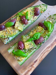 Two Egg Salad Cucumber Boats topped with sliced avocado, Kalamata olives, fresh basil leaves, hemp seeds, and seasoning, served on a wooden board against a dark background.