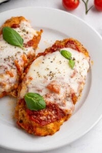 A plate with two pieces of chicken parmigiana topped with melted cheese, tomato sauce, and fresh basil leaves. There are whole cherry tomatoes in the background.