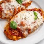 Two breaded chicken parmigiana covered in melted mozzarella cheese and tomato sauce, garnished with fresh basil leaves, served on a white plate.