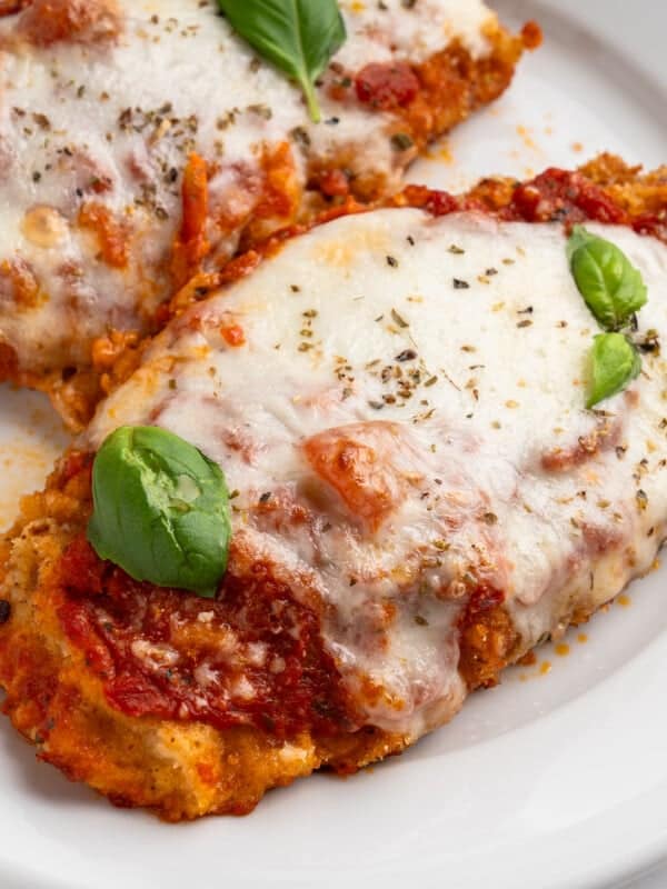 Two breaded chicken parmigiana covered in melted mozzarella cheese and tomato sauce, garnished with fresh basil leaves, served on a white plate.