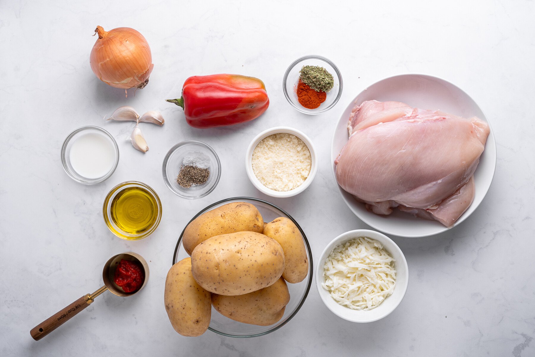 Overhead view of raw ingredients for Chicken and Mashed Potatoes on a white surface: potatoes, chicken breasts, red bell pepper, onion, garlic, shredded cheese, olive oil, milk, spices, and tomato paste in small bowls. - 4
